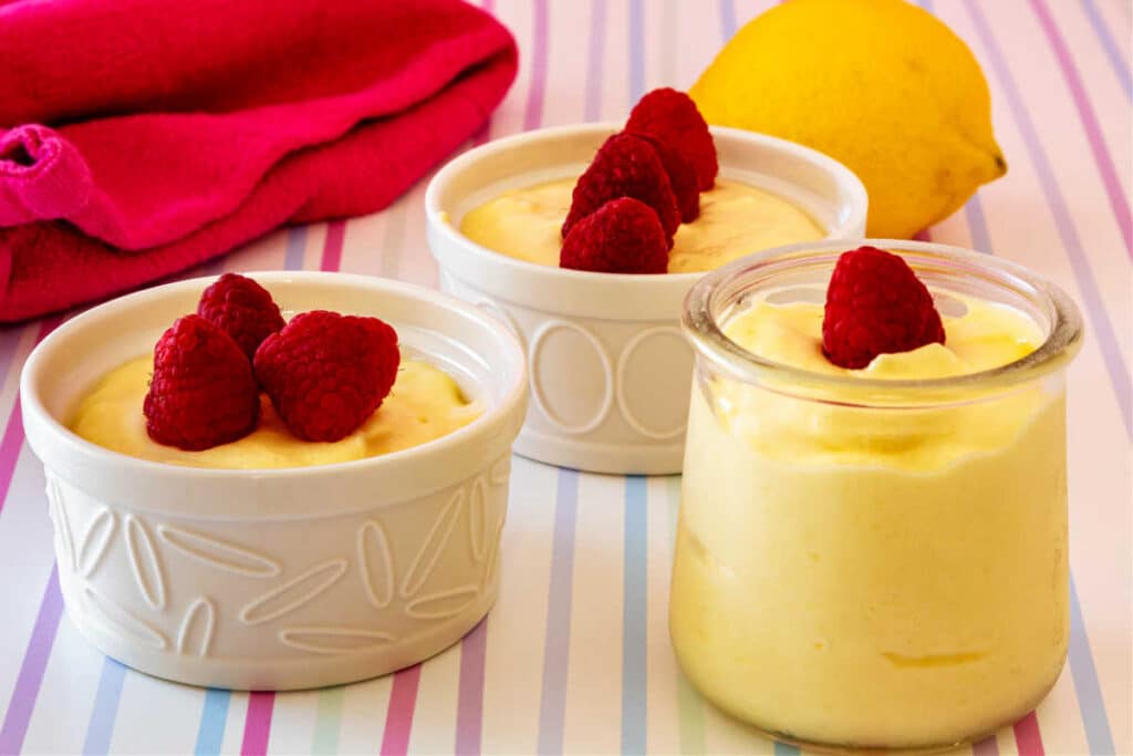 Two white ramekins and one small glass jar of lemon mousse all topped with raspberries.