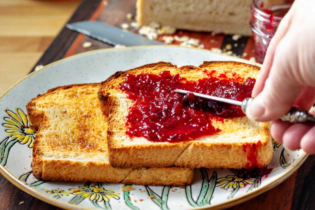 Two slices of toast, overlapped on a round plate. A hand is spreading bright red jam on the top slice.