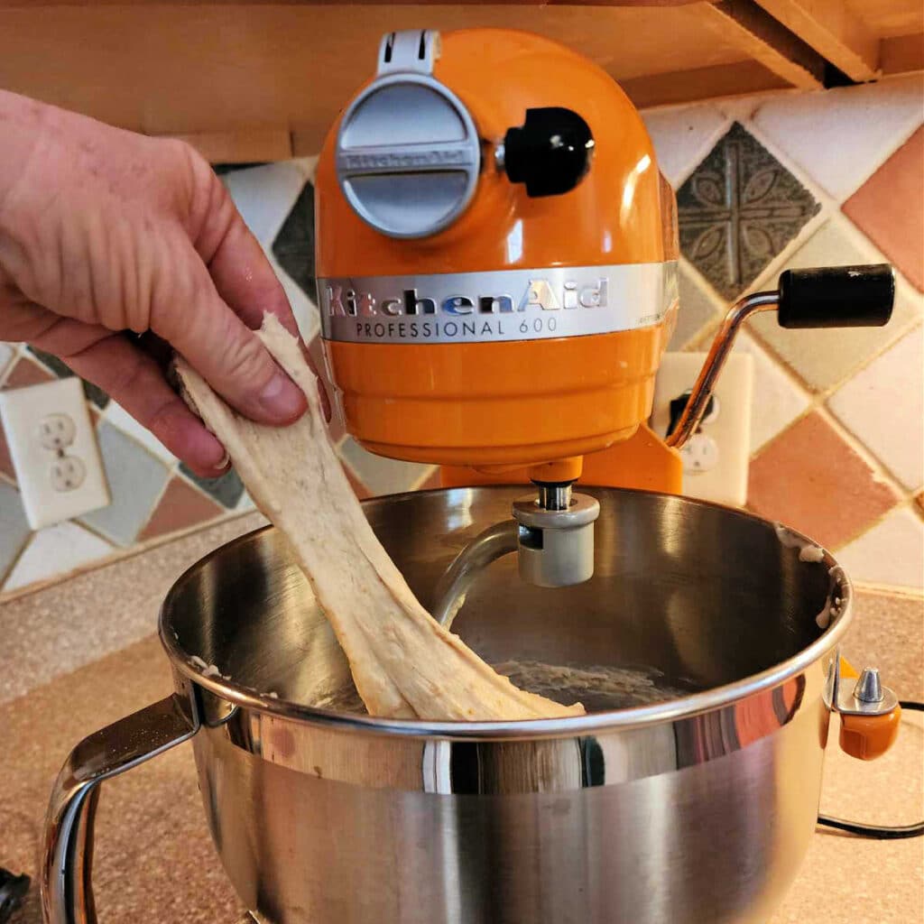 A hand pulling fully-kneaded dough out of the bowl of an orange stand mixer. The dough is very stretchy.