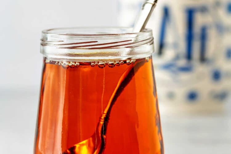 A close-up image of a small, clear glass jar full of caramel syrup with a small silver spoon in it. There's also a coffee mug in the background.