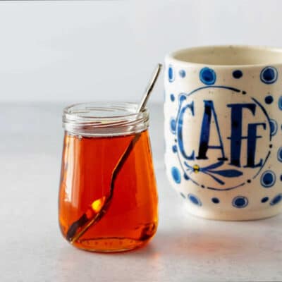 A square image of a small, clear glass jar of amber caramel syrup and a coffee cup.
