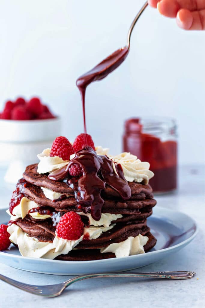 A stack of 5 chocolate pancakes with swirls of whipped cream and raspberries between the layers and on top. A spoon is drizzling chocolate raspberry sauce over the top of the stack.