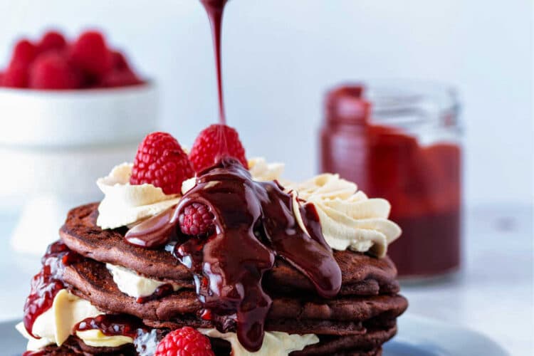 A stack of 5 chocolate pancakes with swirls of whipped cream and raspberries between the layers and on top. A spoon is drizzling chocolate raspberry sauce over the top of the stack.