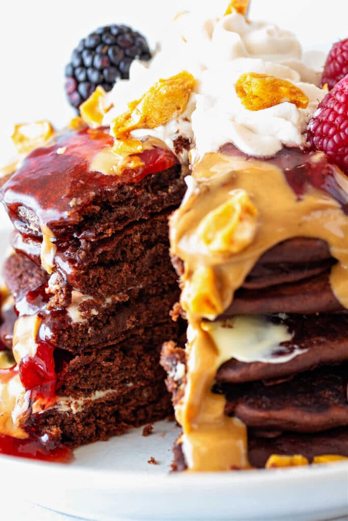 A close-up of a stack of chocolate pancakes with strawberry sauce and peanut butter sauce poured over them. The stack is cut so a wedge is missing showing the fluffy, tender crumb of the pancakes.