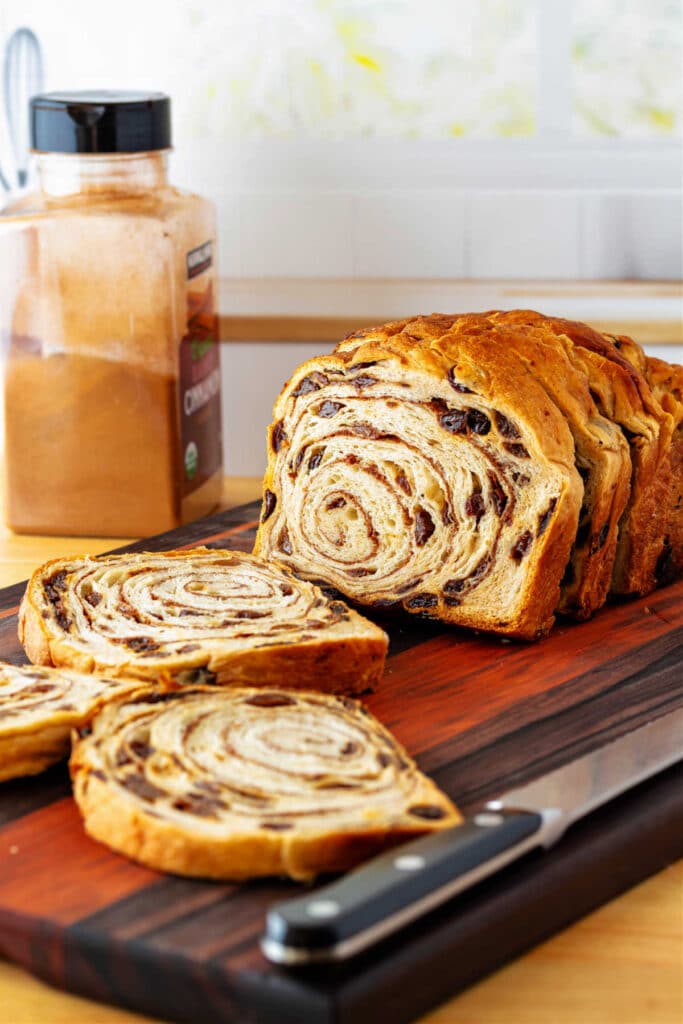 A sliced loaf of cinnamon raisin bread on a cutting board with a bread knife and a jar of ground cinnamon in the background.