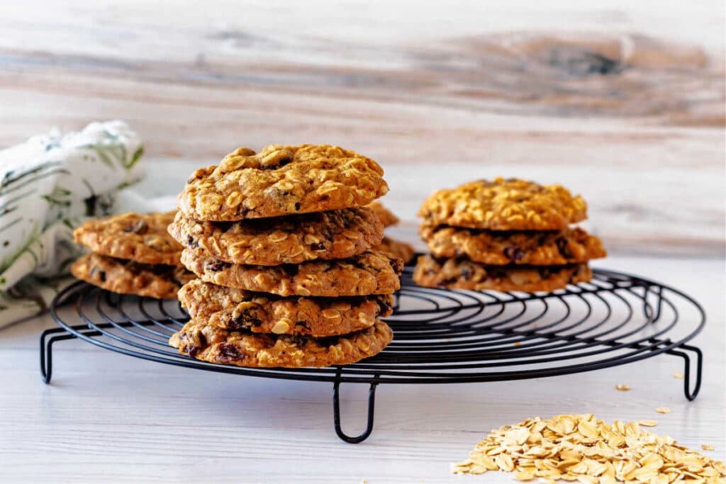 A horizontal image of two stacks of cookies on a cooling rack with some rolled oats in the foreground and a white and green print napkin in the background.