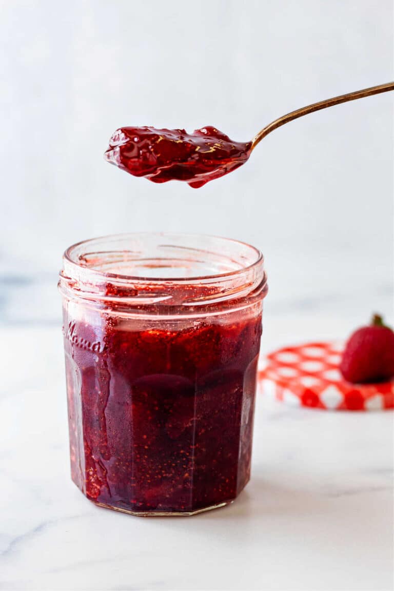 A glass jar full of ruby red strawberry preserves with a spoon of preserves suspended over the jar.