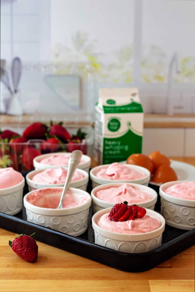 Ramekins of pink, strawberry mousse on a tray in a kitchen with a container of strawberries and a carton of heavy cream in the background.