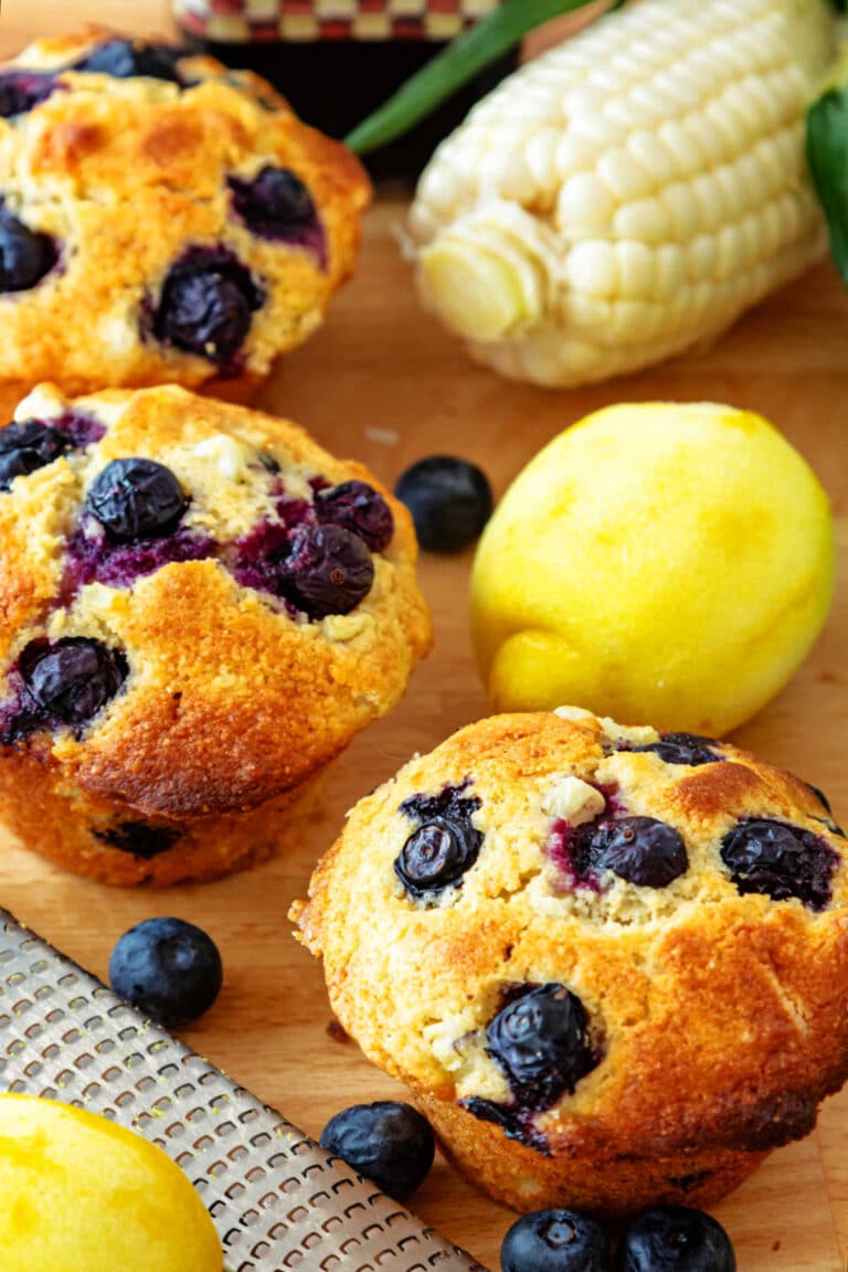 A high-angle shot of 3 corn muffins studded with blueberries along with 2 lemons, an ear of corn, and some blueberries scattered around.