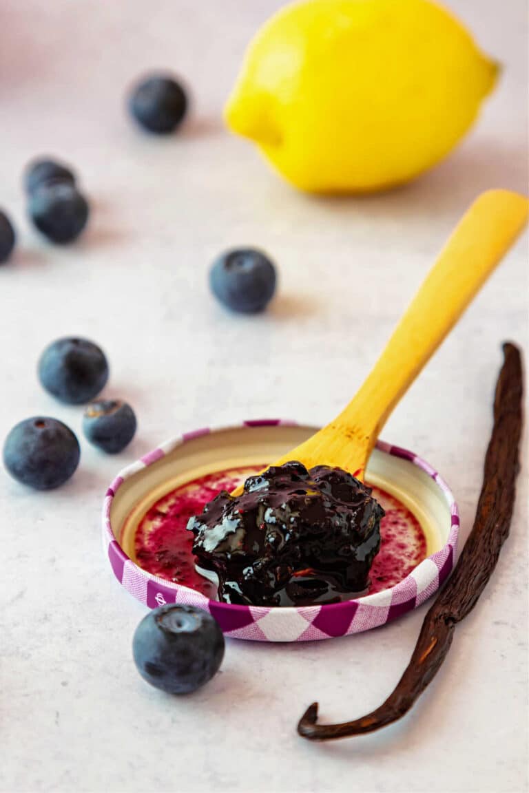 A vertical image showing a jar lid with a wooden spoon resting on it. The spoon is overflowing with blueberry vanilla jam, and there is a vanilla bean, several blueberries, and a lemon in the background.