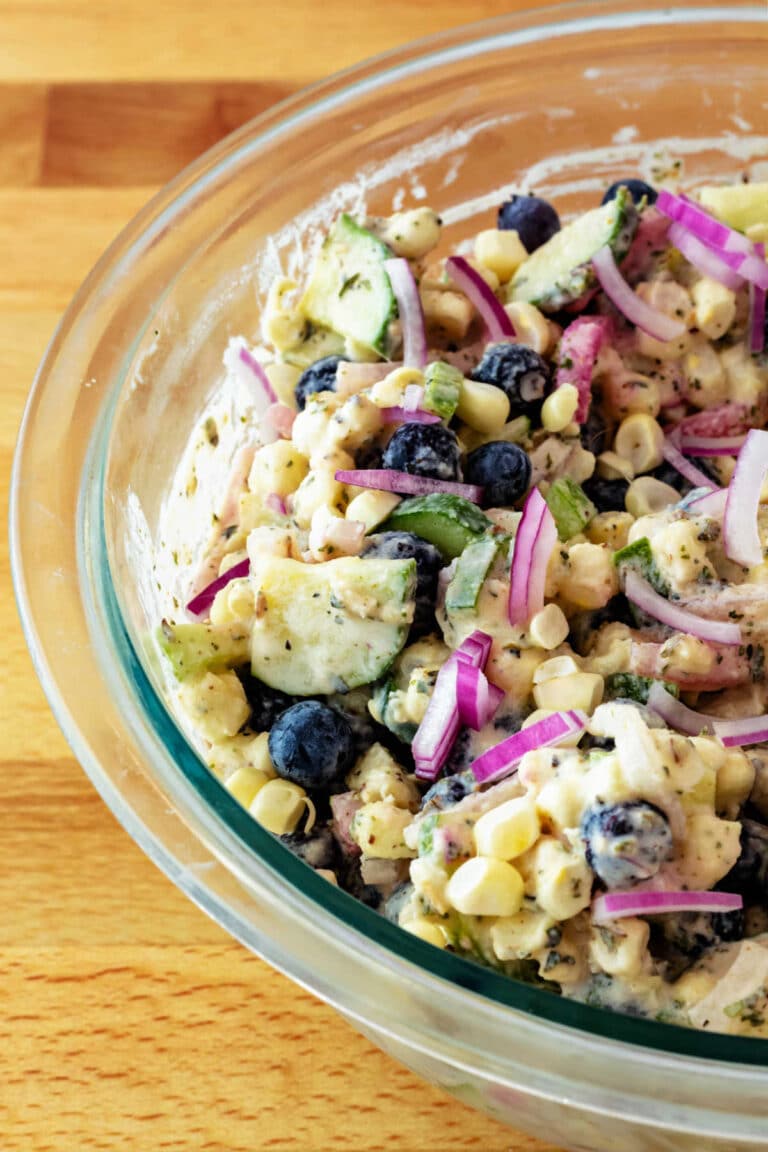 A high-angle shot into a glass bowl of a creamy salad with red onion, white corn, blueberries, and cucumbers.