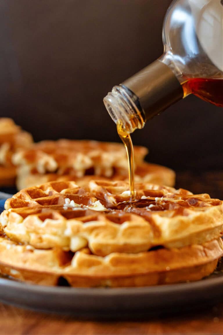 A close-up of maple syrup pouring on a short stack of overnight malted waffles on a brown plate.