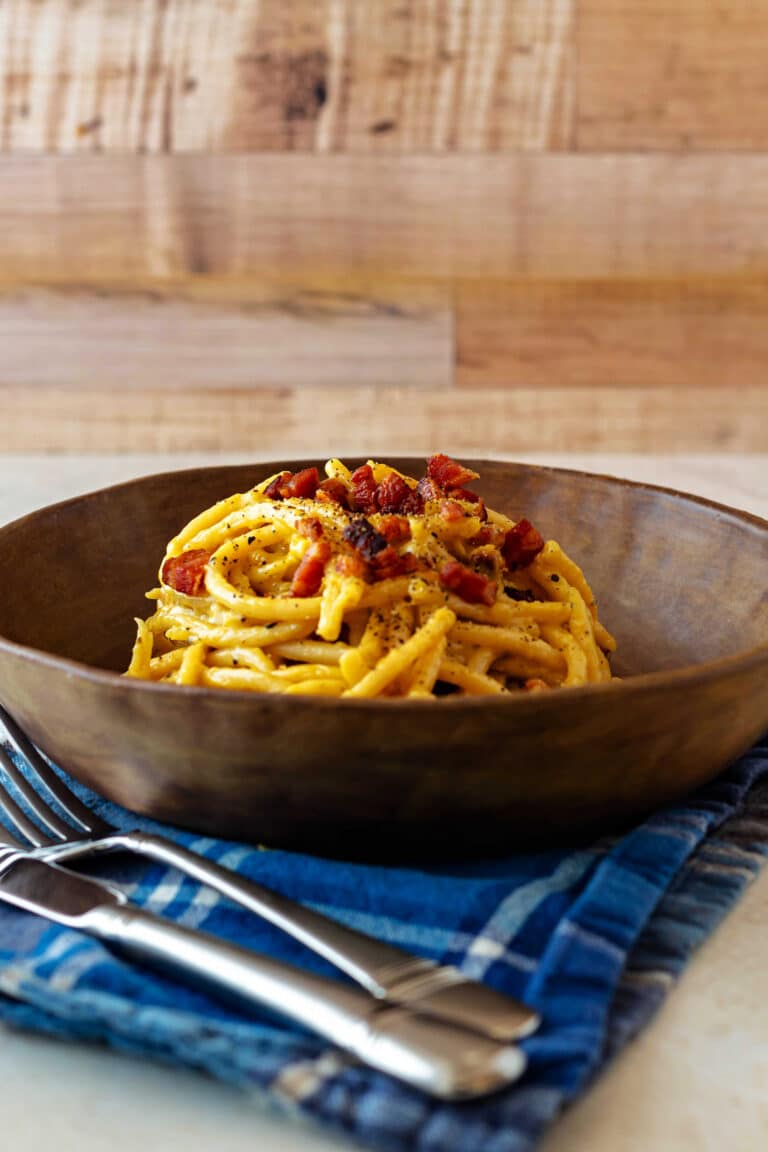 A bowl of bucatini carbonara topped with crispy bacon and black pepper on a blue napkin with a knife and fork in the foreground.