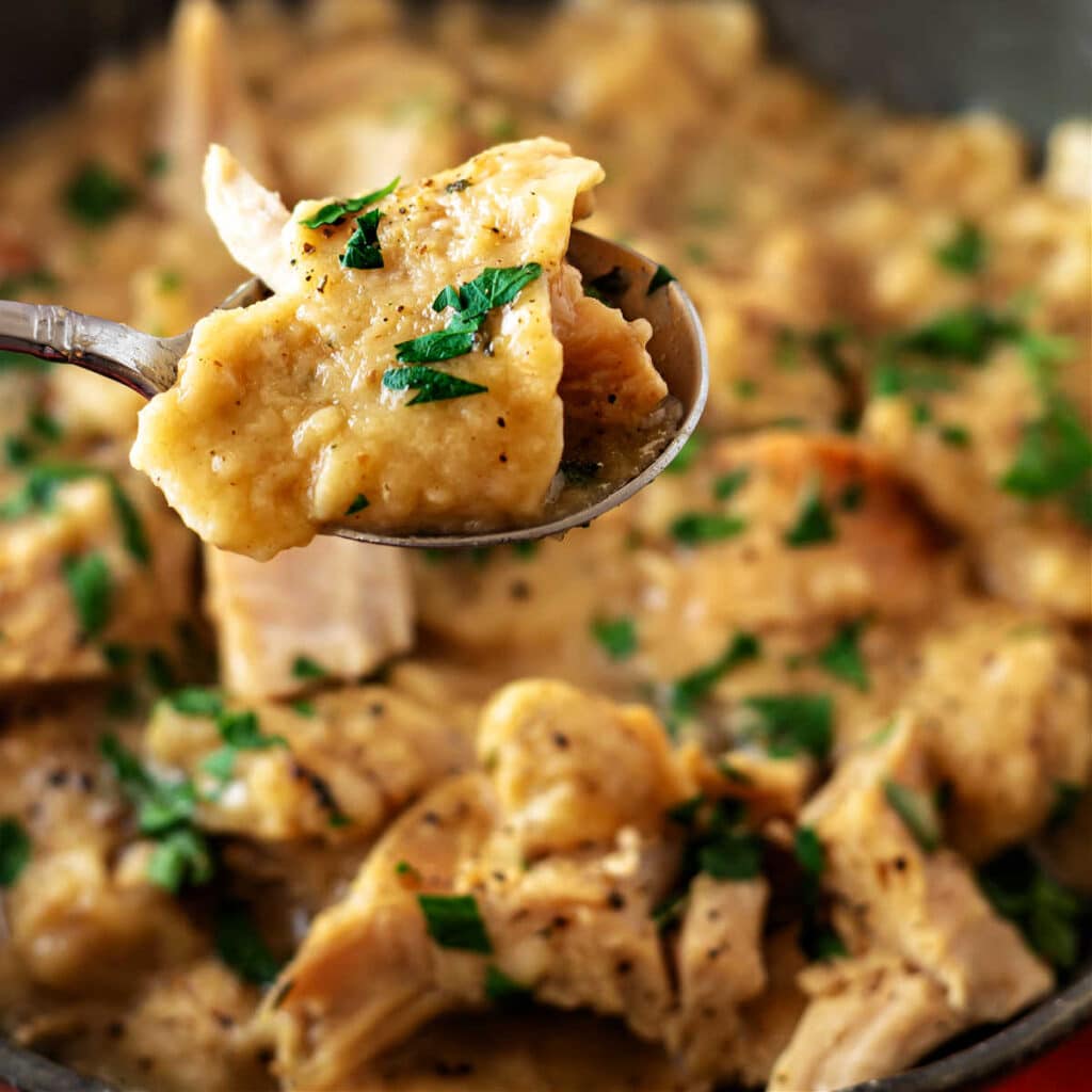 A very close-up shot of a spoon with a bite of turkey and a flat dumpling made of stuffing held over an out-of-focus bowl of more turkey and dumplings.
