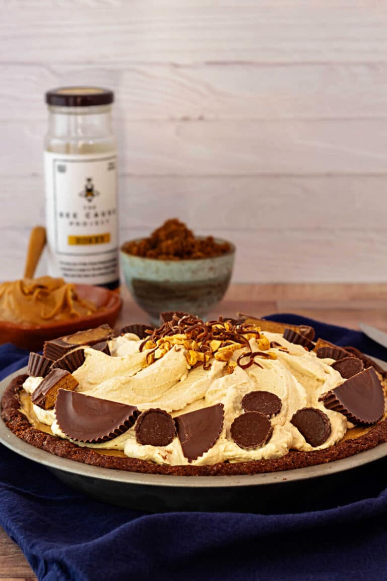 I whole peanut butter cloud pie in a chocolate sable crust decorated with halved and quartered peanut butter cups, chopped peanuts, and shaved chocolate. In the background are dishes of peanut butter, dark brown sugar,a nd a jar of honey.