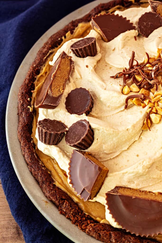 A partial view of peanut butter mousse pie in the pan, showing chocolate crust, creamy filling, whipped topping, peanut butter cups, and chopped peanuts.