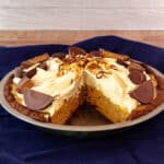 A bountiful peanut butter pie in a chocolate crust with one slice cut from it. The pie is decorated with half and quartered peanut butter cups, mini peanut butter cups, chopped peanuts, and shaved chocolate.