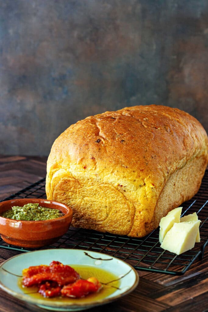 An uncut loaf of bread on a cooling rack surrounded by a dish of sun-dried tomatoes, a small dish of basil pesto, and some small chunks of Parmesan cheese.