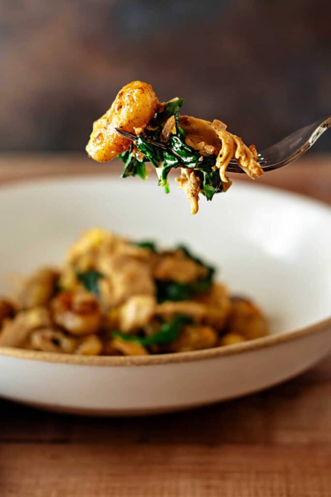 Fork holding a bite of golden pan-roasted gnocchi with lemon cream chicken and spinach above a shallow bowl.