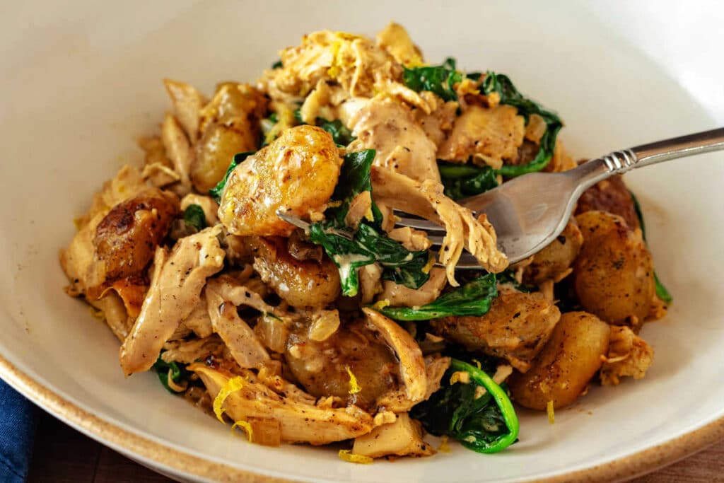 Close-up of golden pan-roasted gnocchi with lemon cream chicken and spinach with a fork lifting a bite from the bowl.