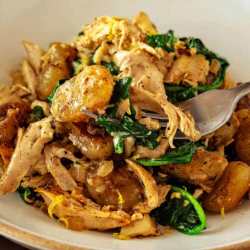 Close-up of golden pan-roasted gnocchi with lemon cream chicken and spinach with a fork lifting a bite from the bowl.