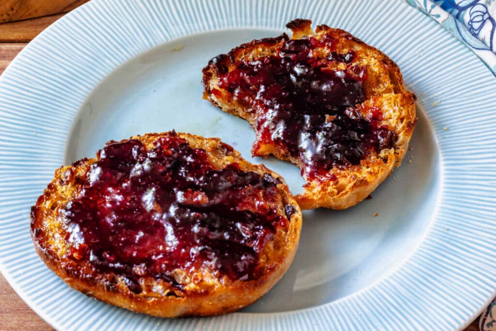 A fork-split and toasted homemade cinnamon raisin English muffin slathered with butter and jam.
