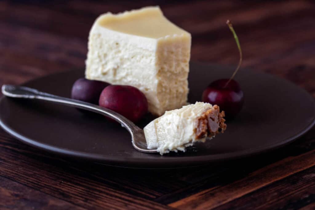 A bite of cheesecake on a fork with the rest of the slice in the background on a brown plate.
