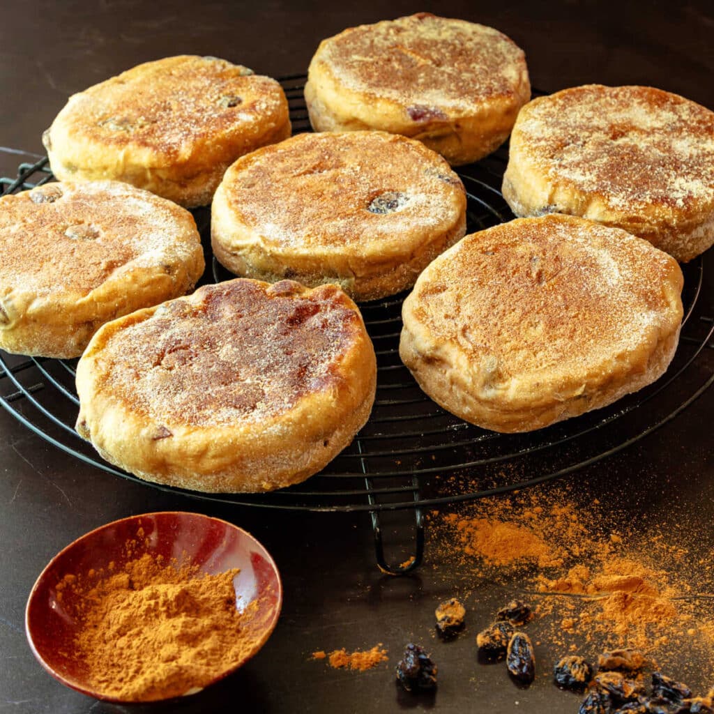 English muffins cooling on a round wire rack with a little bowl of cinnamon and a few raisins scattered in front.