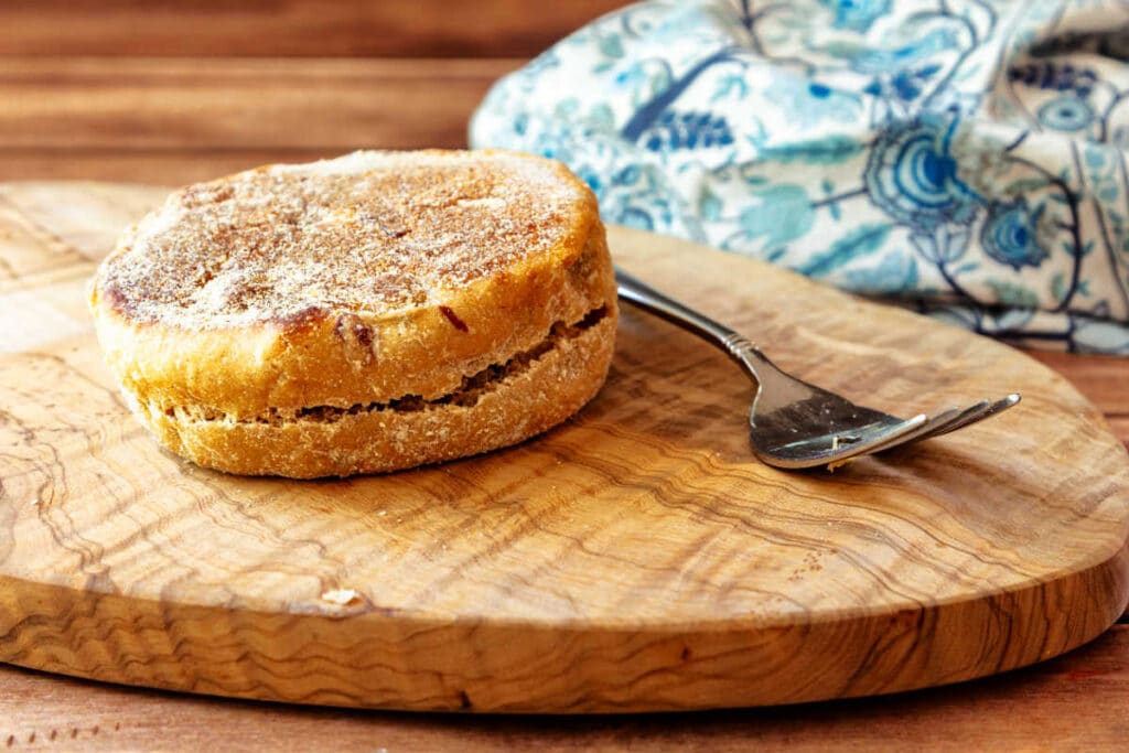 A fork split but not pulled apart English muffin on an olive wood board with a fork next to it.