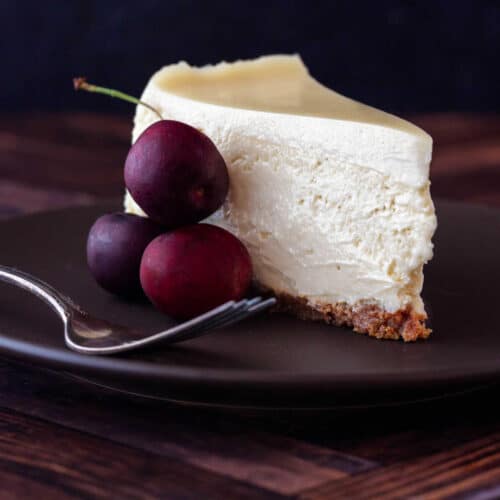 A square image of a cleanly-sliced piece of cheesecake on a brown plate with 3 cherries next to it on the plate.