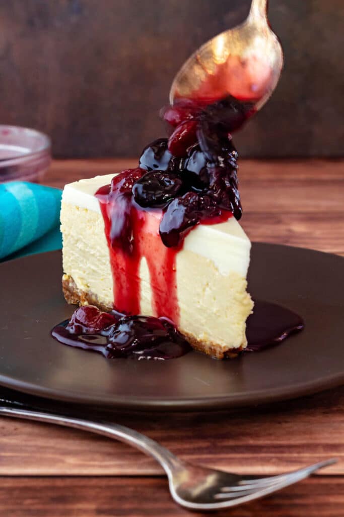 A slice of bakery-style cheesecake with whole cherries in syrup being spooned over the top.