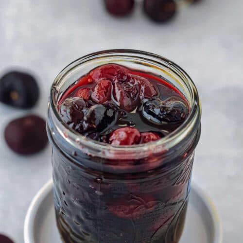 A glass jar of cherry pie preserves with some cherries scattered around.