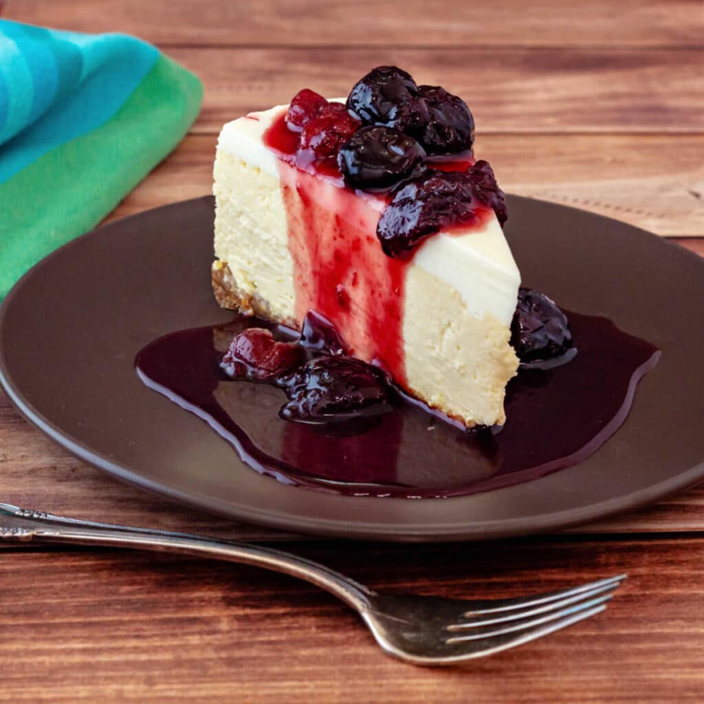 a slice of cheesecake topped with cherry pie preserves. The syrup is running down the sides of the slice and pooling on the plate.