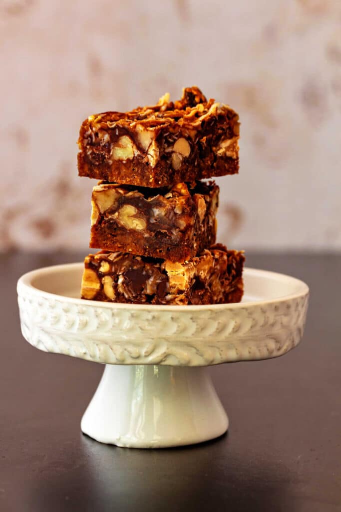A stack of 3 peanut butter butterscotch magic bars on a small, white cupcake pedestal.