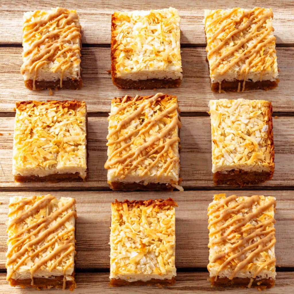 An overhead shot of 9 pb coconut bars arranged in a 3 x 3 grid. Some have a peanut butter drizzle on top while others are just plain lightly toasted coconut.