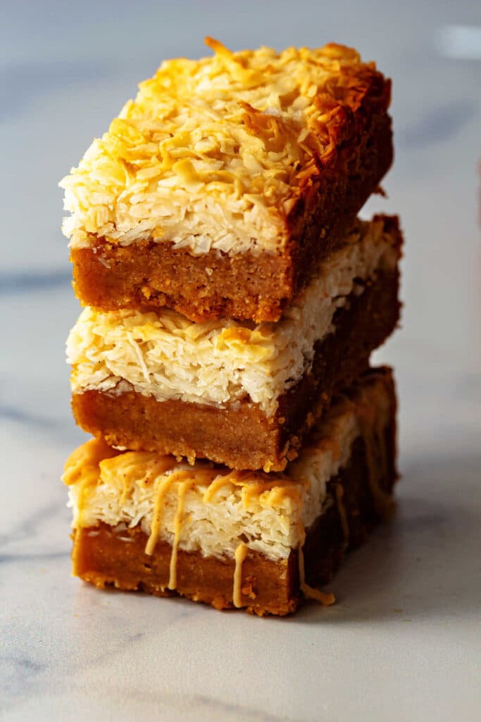 Three stacked peanut butter coconut bars showing equal layers of peanut butter dough and macaroon dough, the fudgy peanut butter interior, and the chewy coconut layer.