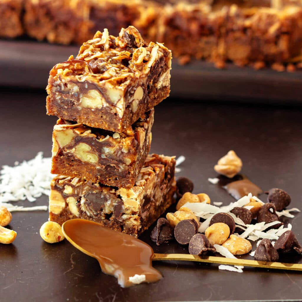 A stack of three 7-layer bars surrounded by some of the mix-in ingredients: coconut, peanut butter chips, peanuts, butterscotch sauce, and chocolate chips.