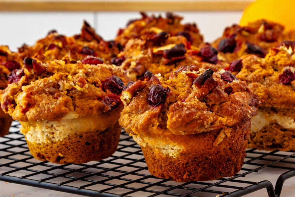 Pumpkin muffins with a Greek yogurt swirl cooling on a rack. From the sides, you can see the ribbon running through the muffin. They are studded with cranberries and chopped pecans.