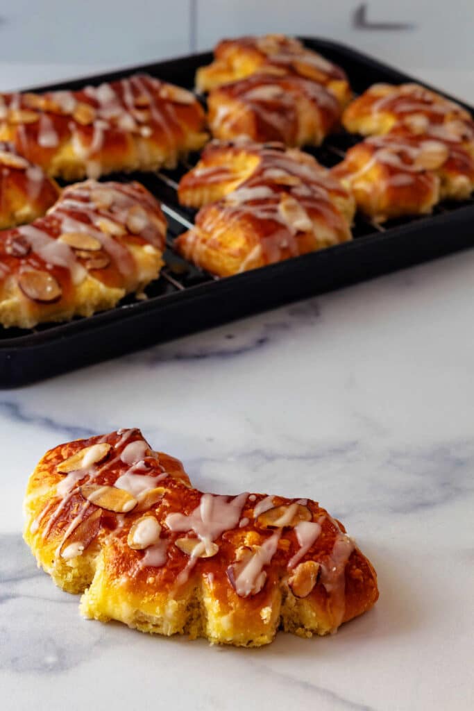One bear claw topped with sliced almonds and a light drizzle of glaze on a marble countertop. The rest of the batch of bear claws are on a black tray in the background.