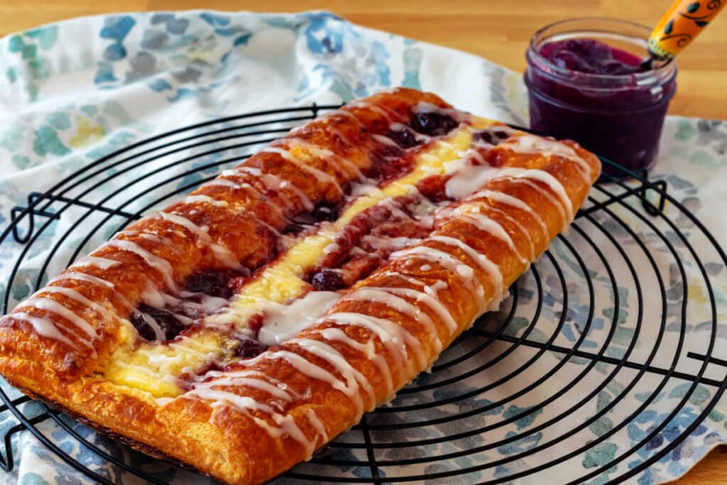 A whole fruit and cheese Danish drizzled with glaze on a round, black wire cooling rack with a small jar of jam in the background.