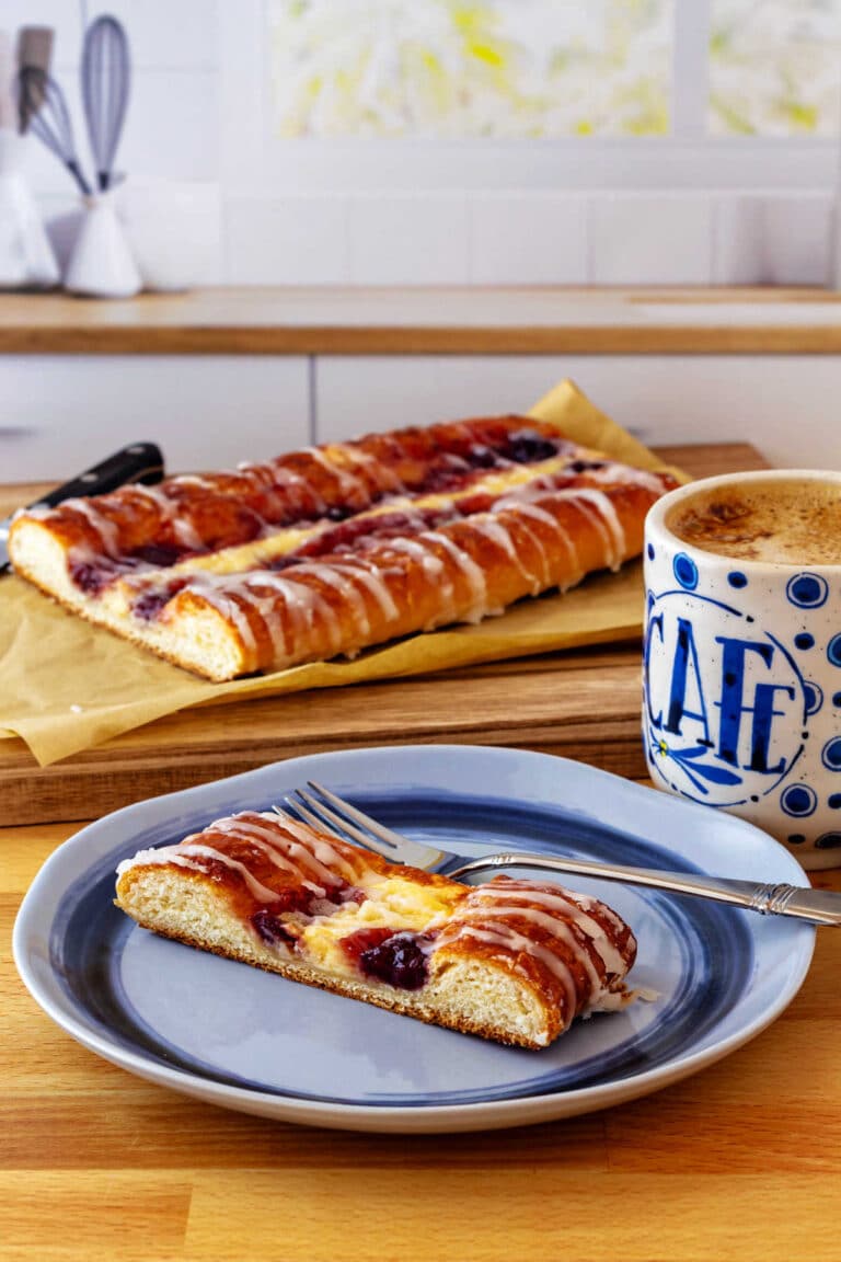 A slice of cheese and fruit Danish on a plate with a cup of coffee and the rest of the Danish on a cutting board in the background.