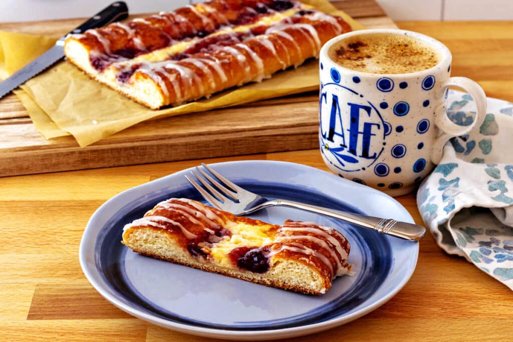 A slice of cheese and fruit Danish on a blue plate with the rest of the Danish in the background.