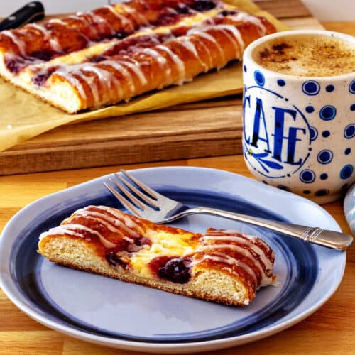 A slice of cheese and fruit Danish on a blue plate with the rest of the Danish in the background.