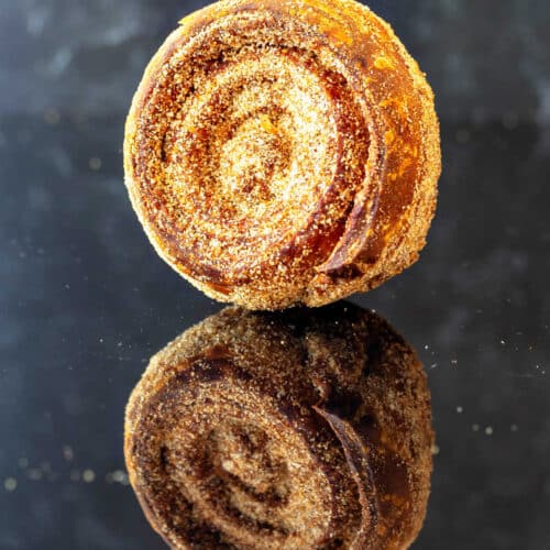 One baked morning bun coated in granulated sugar with the coils toward the camera. It is on a reflective black surface so the mirror image appears below it.