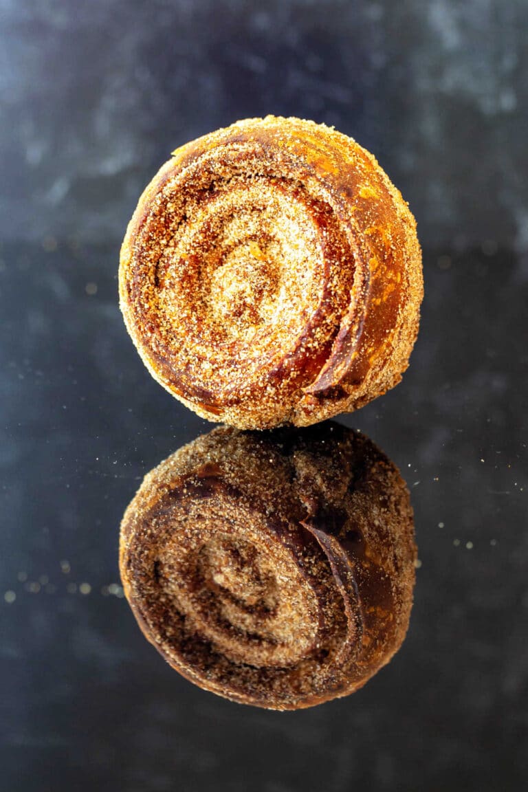 One baked morning bun coated in granulated sugar with the coils toward the camera. It is on a reflective black surface so the mirror image appears below it.