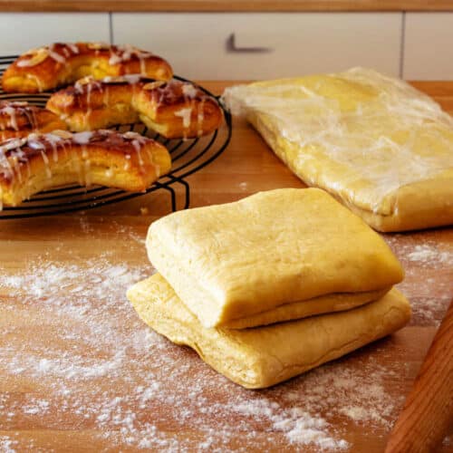 Laminated dough on a floured countertop with a rolling pin and baked bear claws in the background.