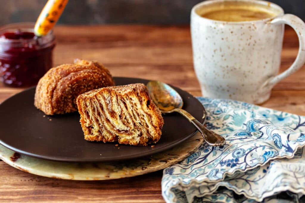 A cut morning bun on a brown plate with a cup of coffee and a jar of jam in the background.
