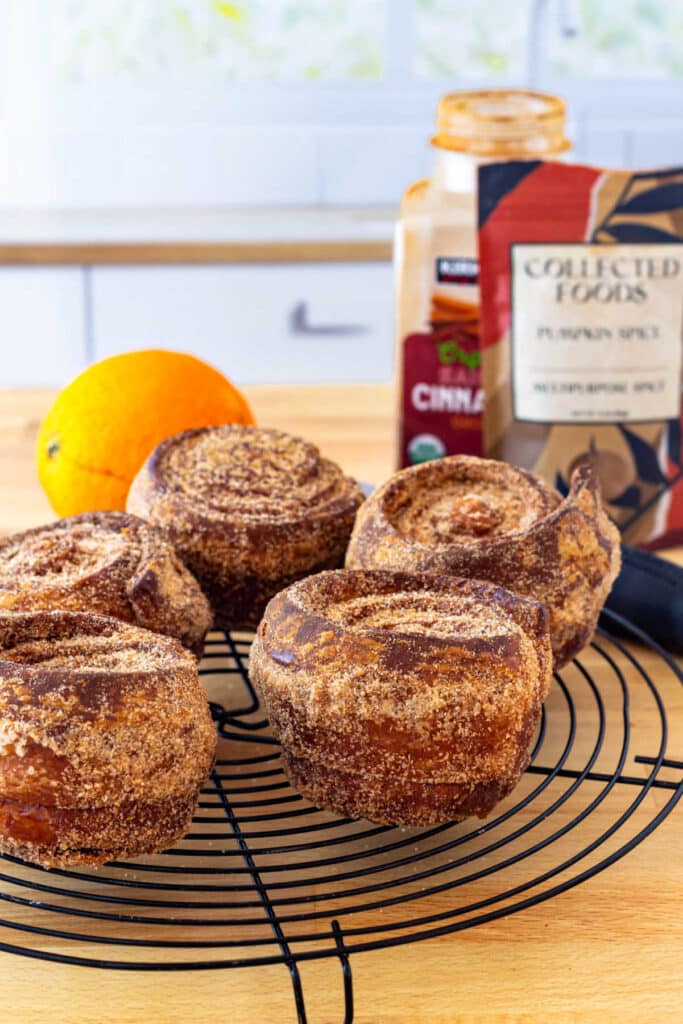 Several large, sugar dusted morning buns cooling on a round wire rack with an orange, a jar of cinnamon, and a packet of pumpkin spice in the background.